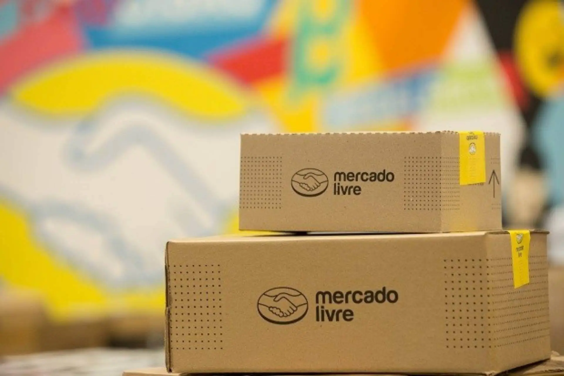 Duas caixas de papelão com logotipos do Mercado Livre estão empilhadas, emolduradas por um fundo colorido atrás delas.