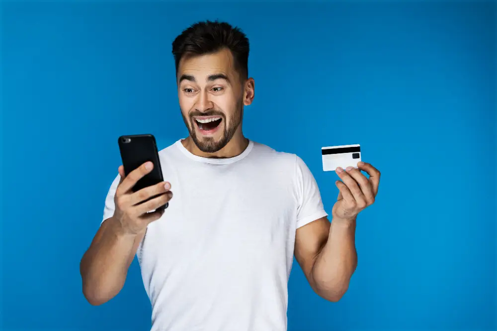 Fundo azul e Homem feliz olhando para seu celular e segurando um cartão de crédito. 