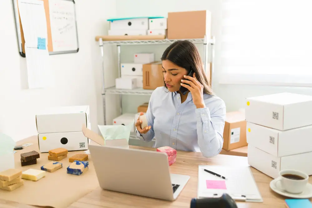 mulher no telefone, sentada em uma mesa com notebook branco sobre a mesa.